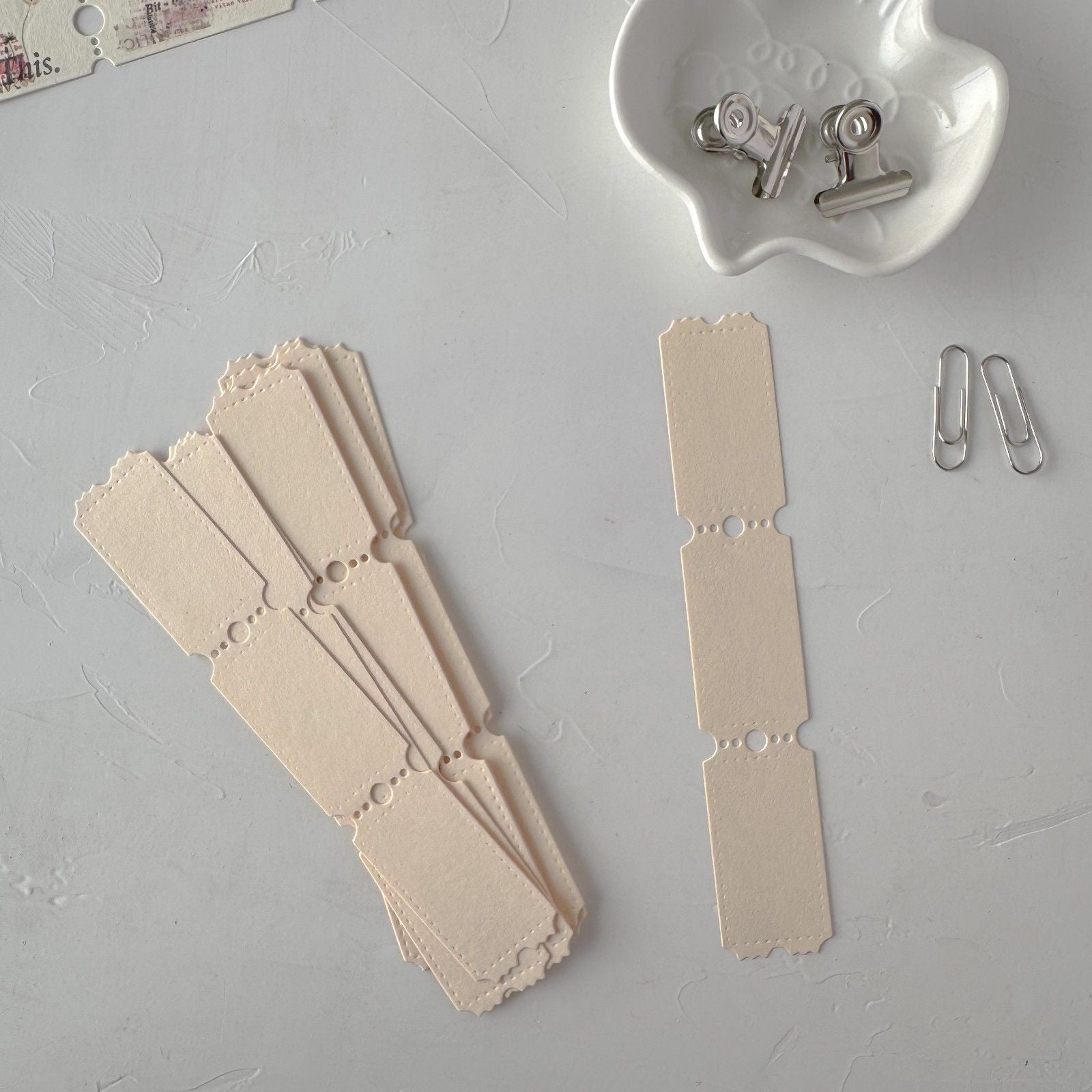 Beige paper tags with decorative edges on a gray surface with metal clips and a white container.