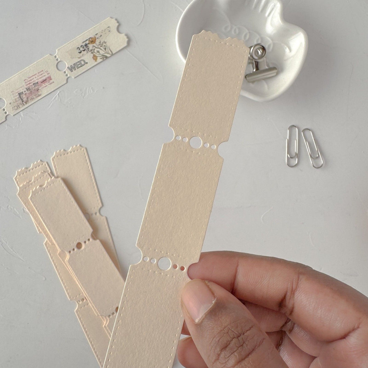 Hand holding up cream Ticket Card Vol. 1 strips with punched holes and small clips