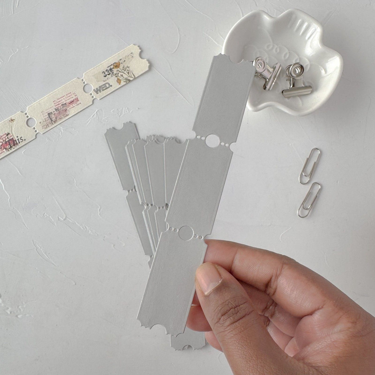 Hand holding a grey Ticket Card Vol. 3 card with more cards and clips on a grey studio surface
