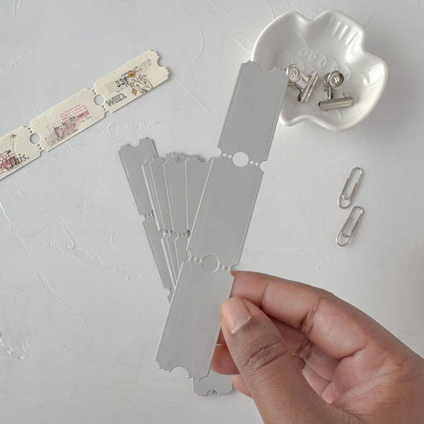Hand holding a grey Ticket Card Vol. 3 card with more cards and clips on a grey studio surface