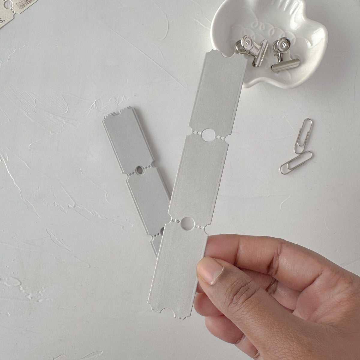 Hand holding a slim grey Ticket Card Vol. 3 strip with small clips on light grey