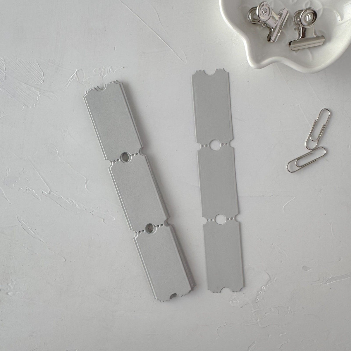 A pair of grey Ticket Card Vol. 3 extended strips with small clips on a grey surface