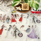 Close-up of decorative PET tape featuring illustrated couples, musicians, and travelers applied on paper, styled with indoor plants and creative journaling accessories.