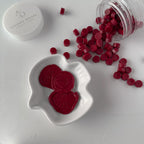 Red wax melts in a heart-shaped dish with an open container of more wax melts on a white background.