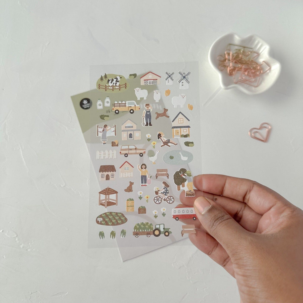 Sheet of illustrated stickers held by a hand beside a dish of paper clips, from the Village Sticker Sheet