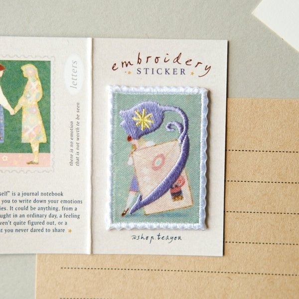 An embroidered sticker card featuring a small stitched patch of a blue cat holding a pink book, with the text 'embroidery STICKER' above it, photographed on a beige paper background.