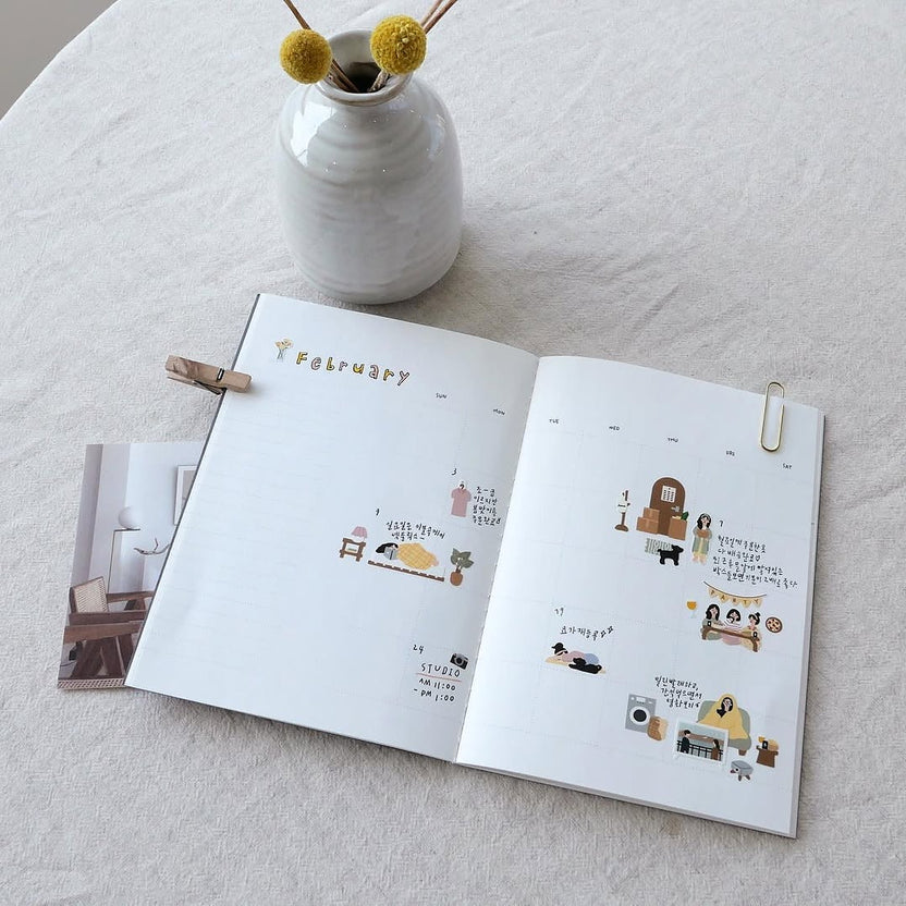 Open book with illustrated pages beside a small vase of yellow flowers, from the Vlog Home Sticker Sheet