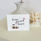 Card with 'home sweet home' text and illustrations on a surface with a vase and decorative items.