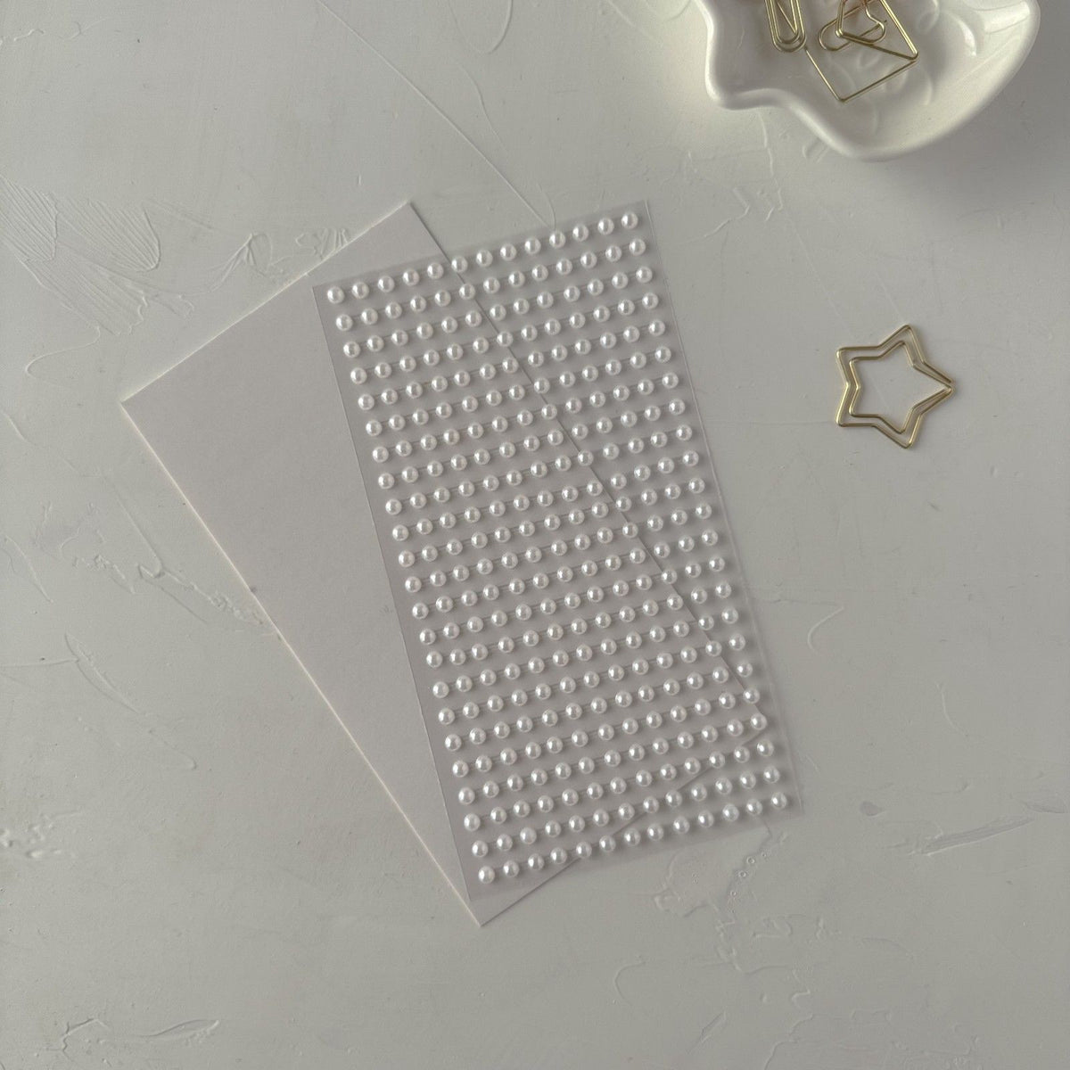 Sheet of medium pearl sticker beads on white beside a star-shaped paper clip, close-up