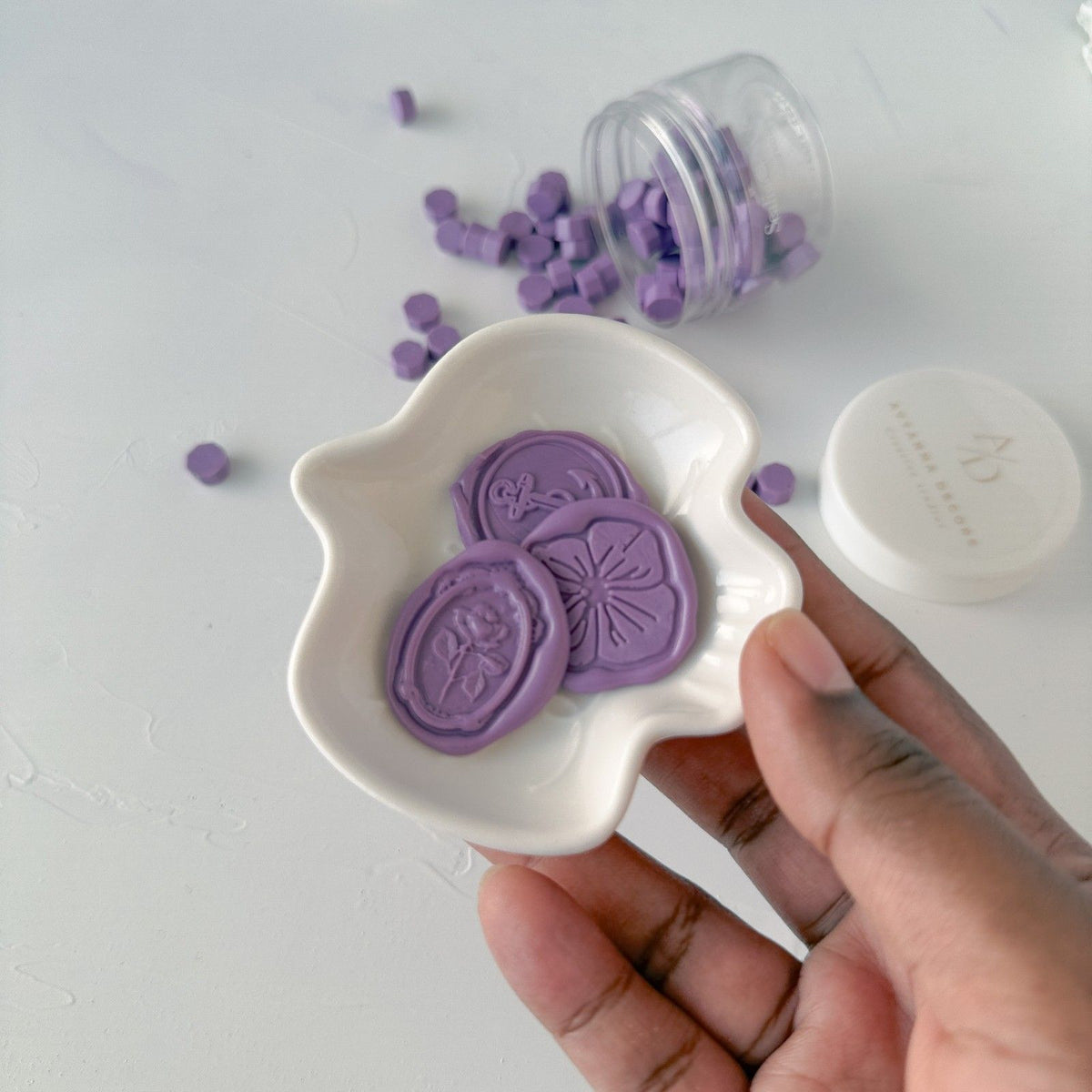 Hand holding a white dish of lavender Wisteria sealing wax beads against a light grey studio backdrop