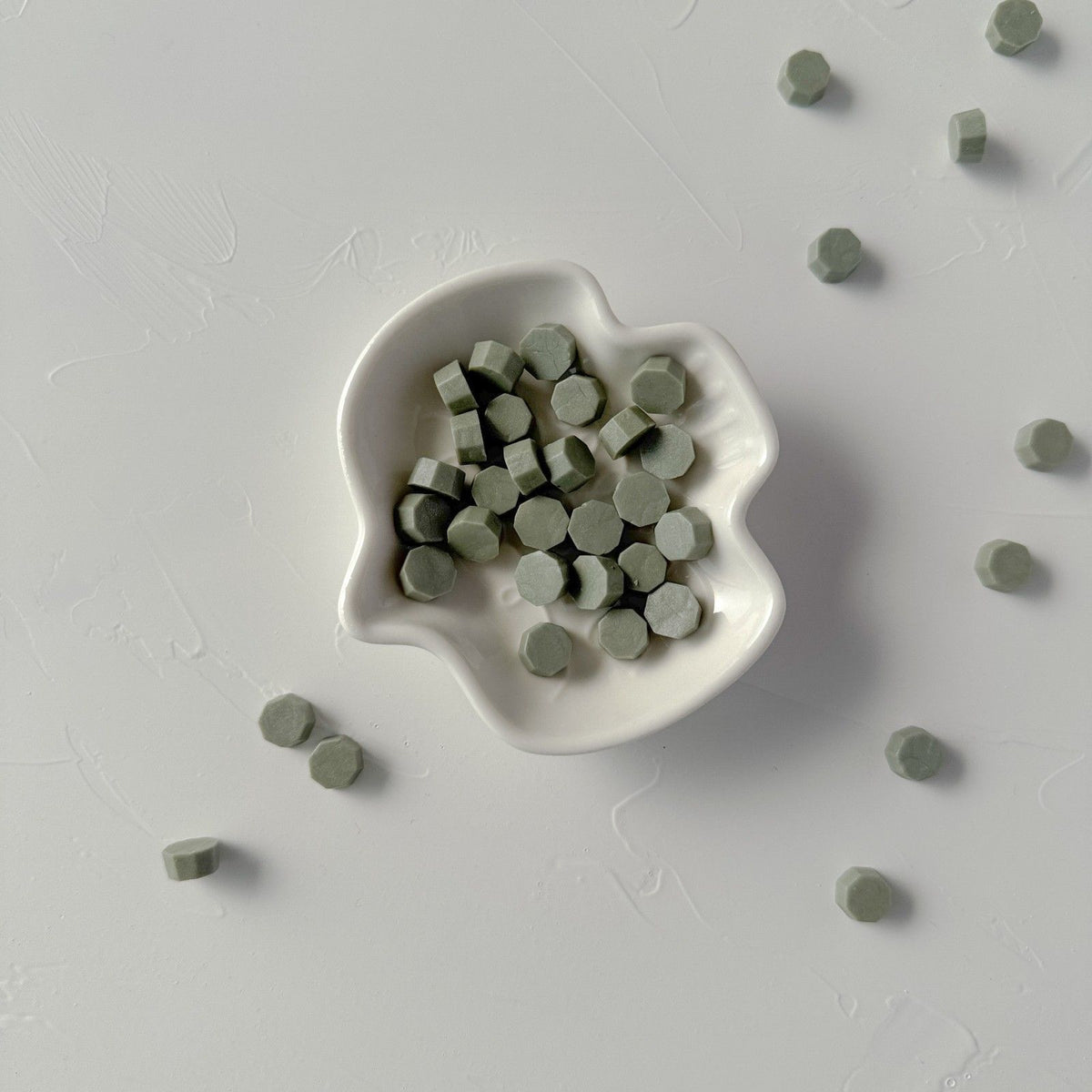 Small white dish of forest-green Woodlands sealing wax beads on a soft grey studio background, top view
