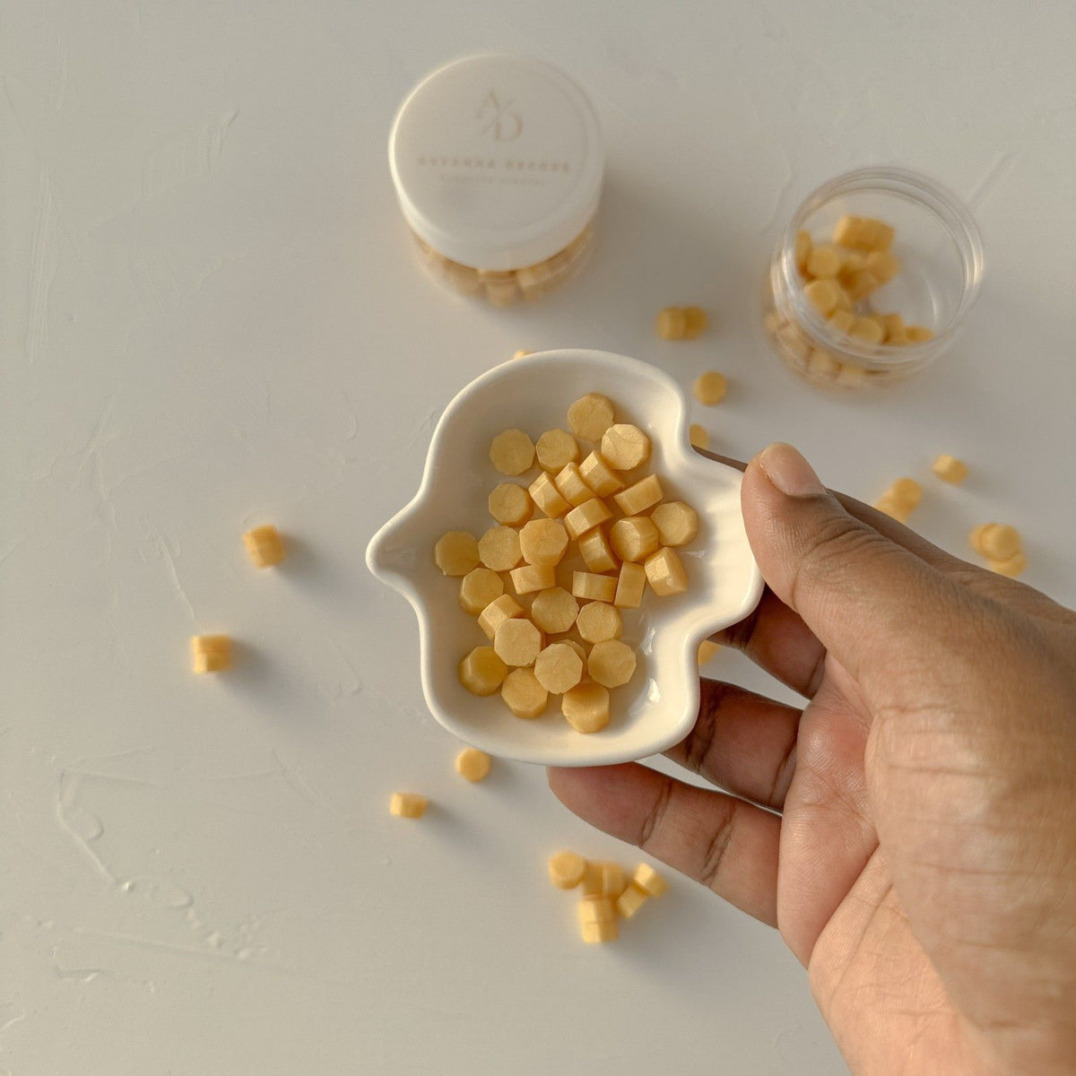 Hand holding a small white dish of bright Yellow Marigold Wax Beads against a light grey background
