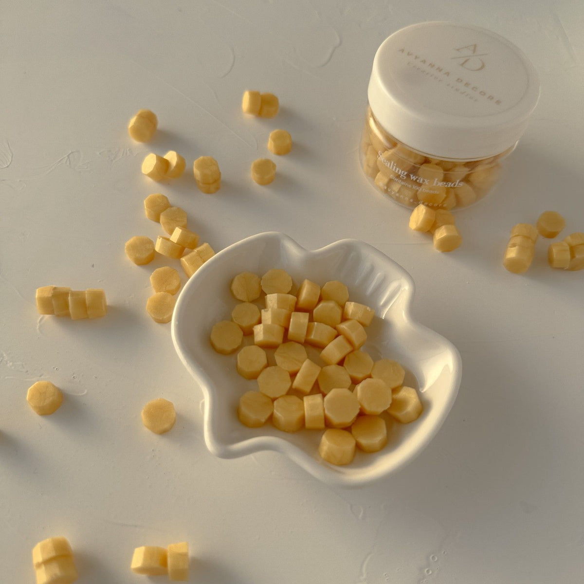 Marigold-yellow wax bead pieces in a white bowl with the labelled jar behind on a soft grey backdrop