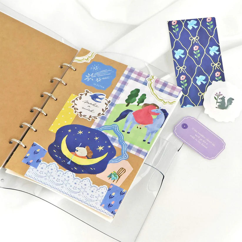 Open ring-bound kraft notebook decorated with colorful stickers, including a moon and stars, a horse illustration, trees, floral patterns, and washi-style borders, arranged on a white surface with matching bookmarks and tags nearby.