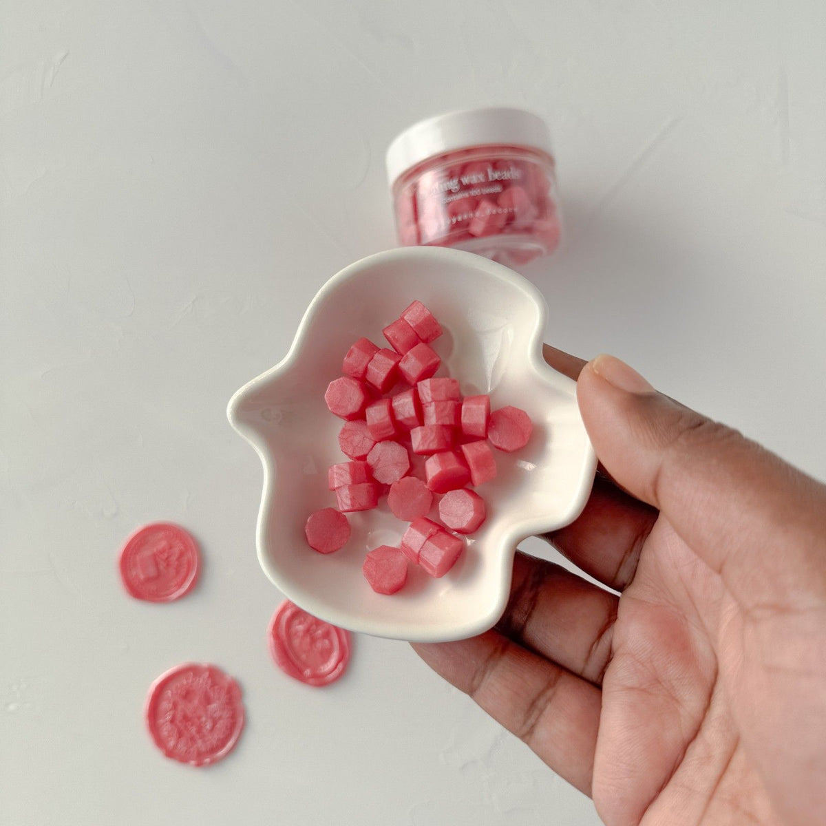 Hand holding a white dish of Young Wine Wax Beads pieces against a light grey studio background