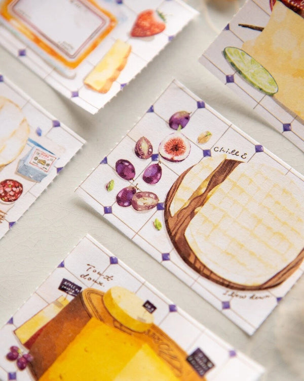 A close-up of several illustrated cheese-themed cards or labels arranged on a light surface. The visible designs include watercolor-style cheese wedges, figs, lime slices, and handwritten labels such as "cheese" and "truffle cheese. - avyanna decore