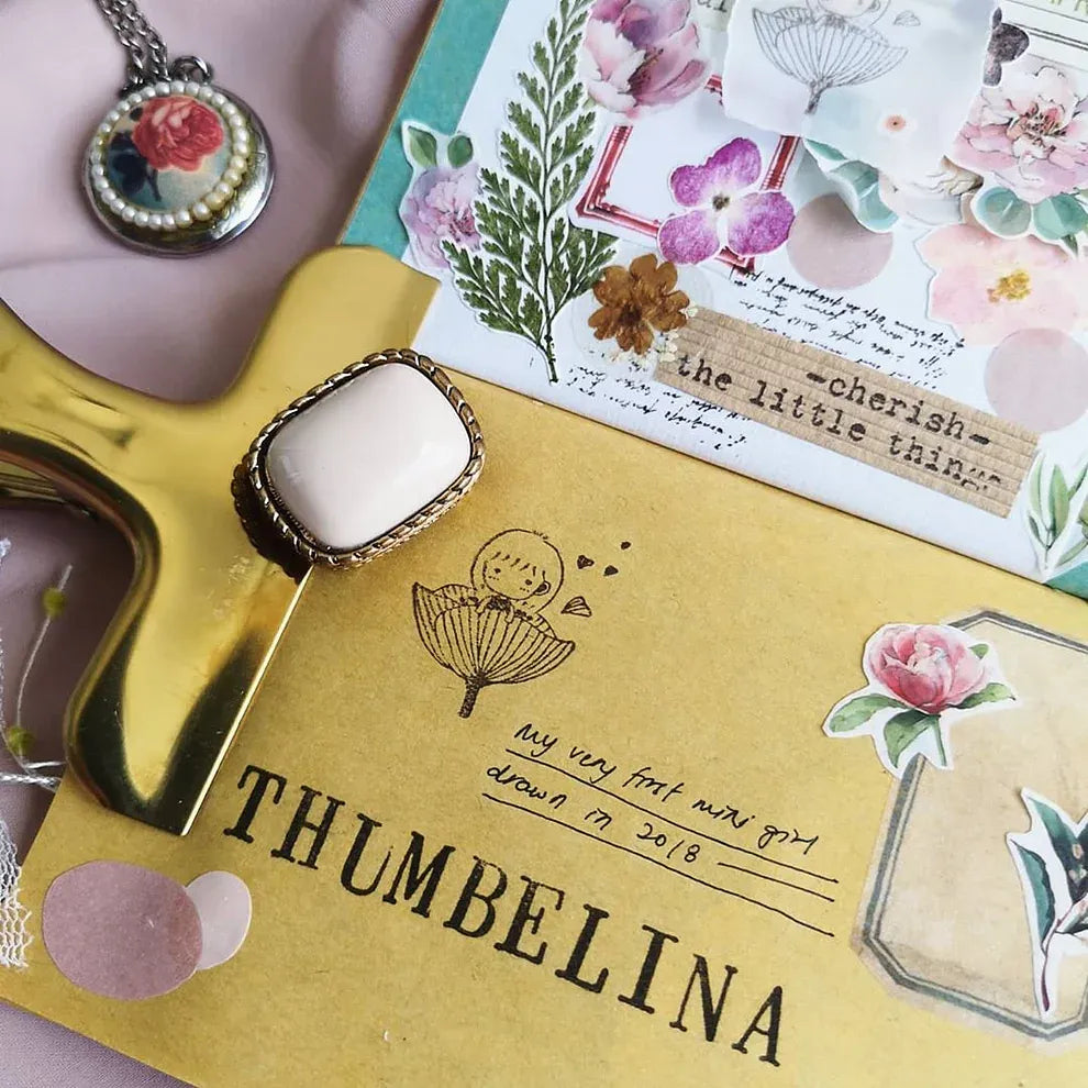 Decorative mirror with 'Thumbelina' design on a yellow surface, surrounded by floral elements and text.