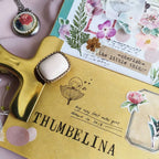 Decorative brooch on a 'Thumbelina' notebook with floral and nature-themed stationery items.