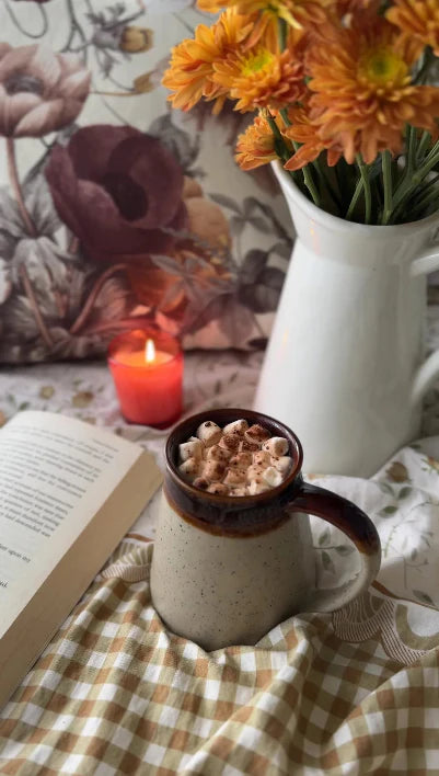Hot chocolate in a mug with whipped cream, a lit candle, and flowers on a checkered tablecloth.