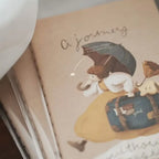 Notebook with a cute illustration of a bear holding an umbrella and a suitcase, with text 'A journey' on a beige background.