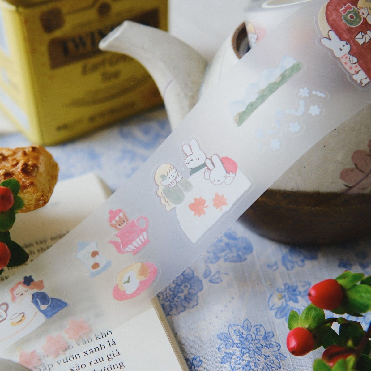 Tea bag with cartoon characters on a floral tablecloth with a teapot and book in the background.