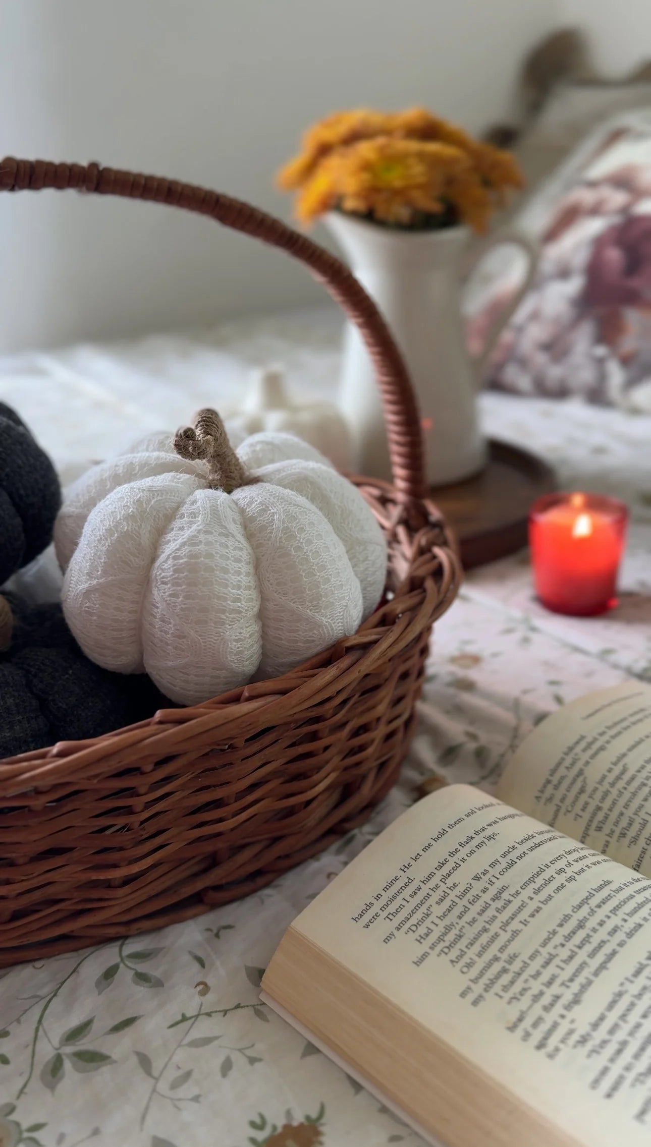 White pumpkin in a wicker basket on a floral-patterned surface with a candle and book in the background.